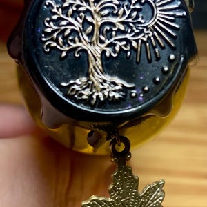 Pode incluir: Um selo de cera preto circular com um design em relevo de uma &aacute;rvore e um sol em dourado. O selo tem um acabamento brilhante com part&iacute;culas de glitter sutis. Um pequeno pingente de folha de bordo em bronze texturizado pende da borda inferior.