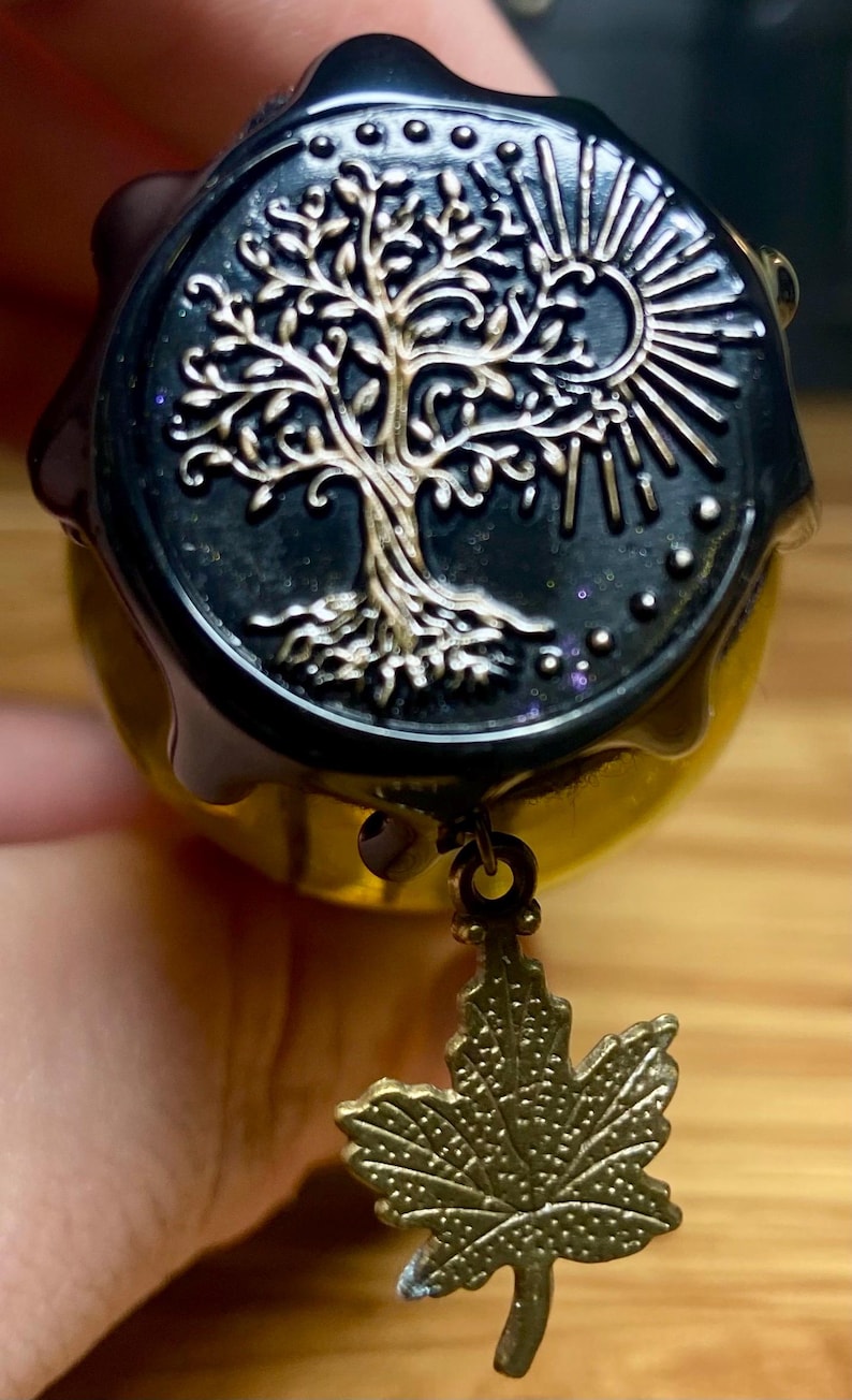 Pode incluir: Um selo de cera preto circular com um design em relevo de uma &aacute;rvore e um sol em dourado. O selo tem um acabamento brilhante com part&iacute;culas de glitter sutis. Um pequeno pingente de folha de bordo em bronze texturizado pende da borda inferior.