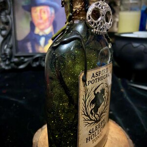 May include: A glass bottle filled with green glittery liquid, topped with black wax drips and a silver skull charm. The label reads ASPIRE APOTHECARY SLEEPY HOLLOW TONIC and features a silhouette of a headless horseman among bare trees.