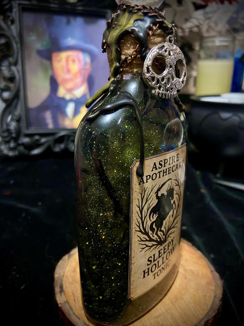 May include: A glass bottle filled with green glittery liquid, topped with black wax drips and a silver skull charm. The label reads ASPIRE APOTHECARY SLEEPY HOLLOW TONIC and features a silhouette of a headless horseman among bare trees.