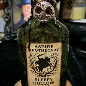 May include: A glass bottle filled with green glittery liquid, featuring a silver skull charm at the neck and black wax drips. The label reads ASPIRE APOTHECARY SLEEPY HOLLOW TONIC and displays a graphic of a headless horseman on a horse.