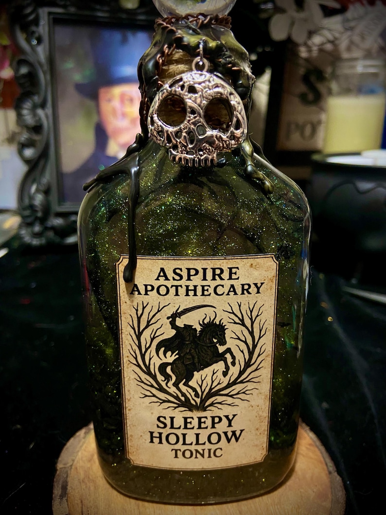 May include: A glass bottle filled with green glittery liquid, featuring a silver skull charm at the neck and black wax drips. The label reads ASPIRE APOTHECARY SLEEPY HOLLOW TONIC and displays a graphic of a headless horseman on a horse.