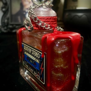 Puede incluir: Una botella de vidrio rectangular con un tap&oacute;n esf&eacute;rico transparente, envuelta en cordel rojo y una cadena plateada. La botella presenta cera roja goteando por los lados y una etiqueta que dice Death By Stereo y The Lost Boys con un casete azul.