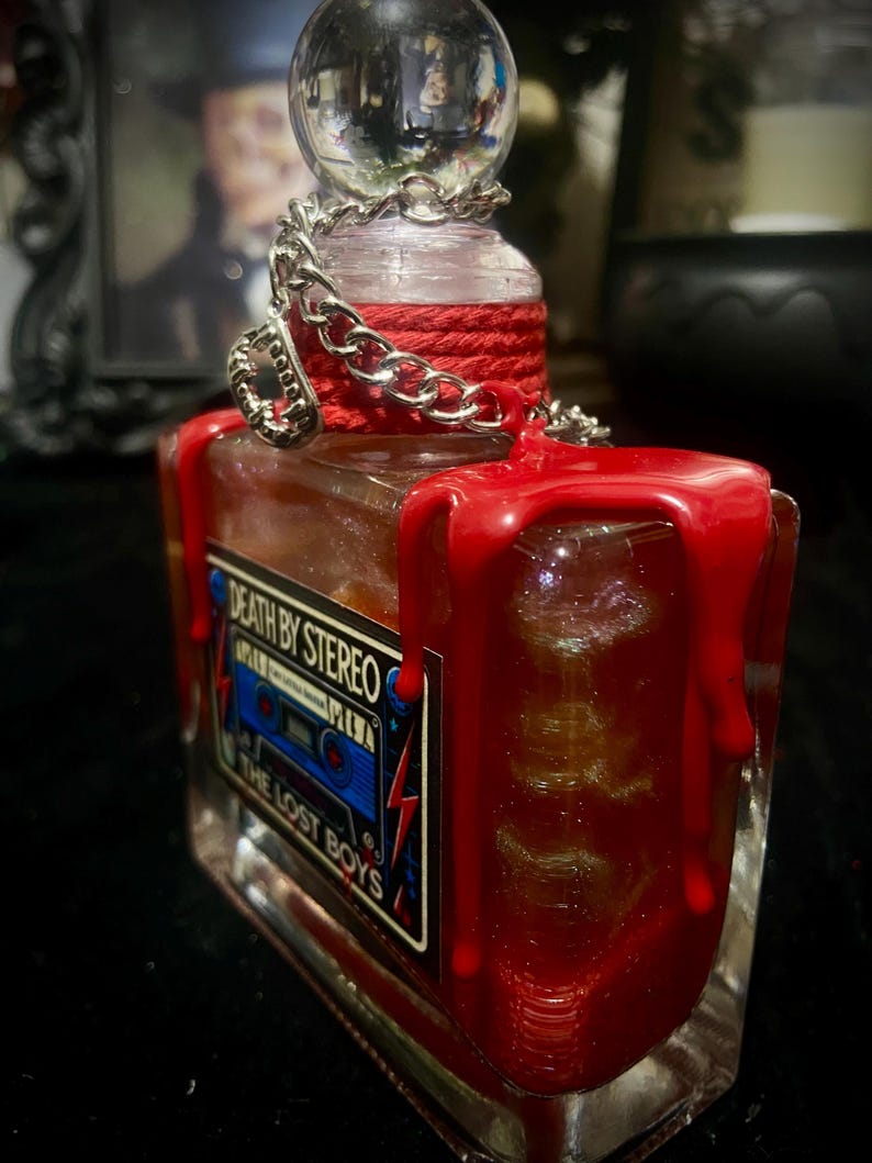 Puede incluir: Una botella de vidrio rectangular con un tap&oacute;n esf&eacute;rico transparente, envuelta en cordel rojo y una cadena plateada. La botella presenta cera roja goteando por los lados y una etiqueta que dice Death By Stereo y The Lost Boys con un casete azul.