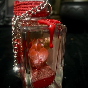 Puede incluir: Una botella de vidrio rectangular transparente con una cuerda roja envuelta alrededor del cuello y una cadena de metal plateado. Dentro hay elementos rojos brillantes y una peque&ntilde;a calavera dorada. Cera roja gotea por la esquina exterior.