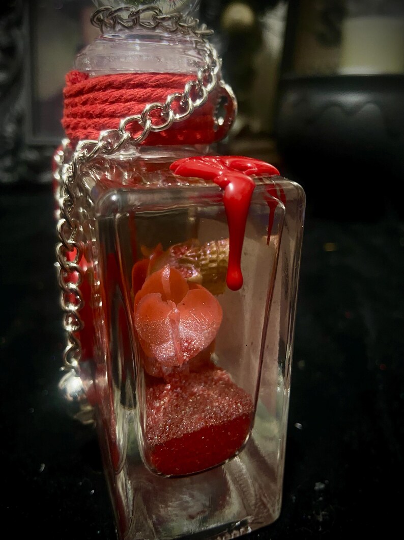 Puede incluir: Una botella de vidrio rectangular transparente con una cuerda roja envuelta alrededor del cuello y una cadena de metal plateado. Dentro hay elementos rojos brillantes y una peque&ntilde;a calavera dorada. Cera roja gotea por la esquina exterior.