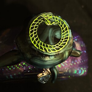 Puede incluir: Un objeto de resina de color morado oscuro y negro que presenta una parte superior circular con un dise&ntilde;o de serpiente ouroboros de color amarillo brillante. La superficie tiene un acabado brillante e iridiscente con purpurina incrustada. Un peque&ntilde;o dije transparente en forma de coraz&oacute;n cuelga del lateral.