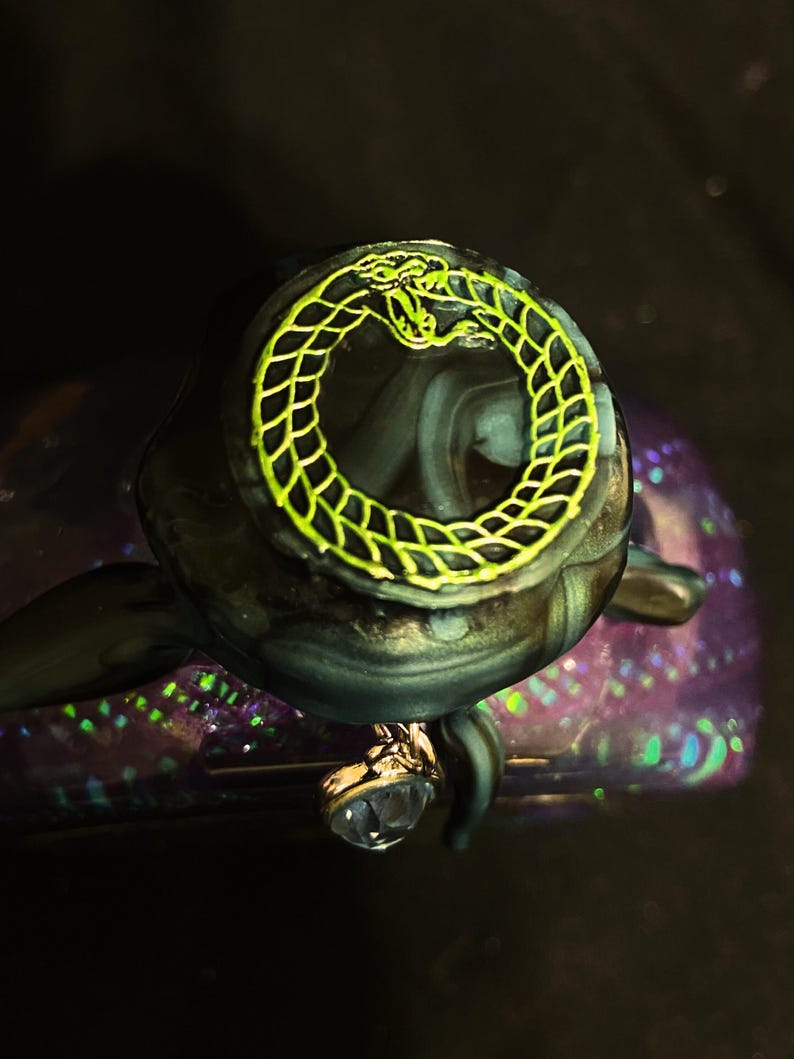 Puede incluir: Un objeto de resina de color morado oscuro y negro que presenta una parte superior circular con un dise&ntilde;o de serpiente ouroboros de color amarillo brillante. La superficie tiene un acabado brillante e iridiscente con purpurina incrustada. Un peque&ntilde;o dije transparente en forma de coraz&oacute;n cuelga del lateral.