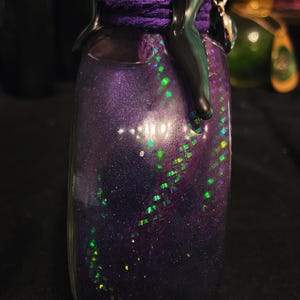 Puede incluir: Una botella de vidrio llena de un l&iacute;quido p&uacute;rpura oscuro brillante que contiene part&iacute;culas de purpurina verde y plateada iridiscente. El cuello est&aacute; envuelto en hilo p&uacute;rpura y cubierto con cera negra que gotea por los lados, con un dije plateado.