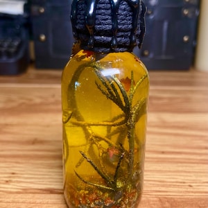 Pode incluir: Uma garrafa de vidro transparente cheia de um l&iacute;quido amarelo dourado, contendo ramos verdes submersos e pequenas part&iacute;culas cor de laranja. O gargalo &eacute; envolvido por um fio preto e selado com cera preta que escorre pelas laterais.