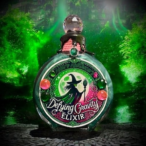 May include: A green glass bottle with a silver glitter finish and a black stopper. The bottle has a label that says "Aspire Apothecary Defying Gravity Elixir" and features a witch with a broom in front of a full moon. The bottle is decorated with pink and green gems.