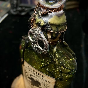 May include: A green glittery glass bottle with a clear spherical stopper, adorned with a silver skull charm, copper-colored chains, and dark wax drips. The label features the text ASPIRE APOTHECARY above a silhouette of a tree and a horse.