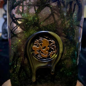 May include: A clear glass bottle filled with dark green liquid, black branch-like structures, and green glitter. A circular wax seal featuring a gold-colored skull and cross motif is applied to the front, with dark green wax dripping down the glass.