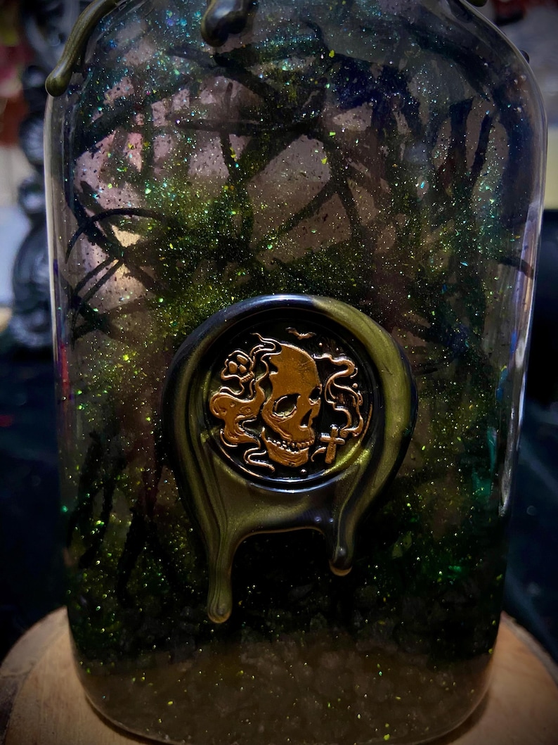 May include: A clear glass bottle filled with dark green liquid, black branch-like structures, and green glitter. A circular wax seal featuring a gold-colored skull and cross motif is applied to the front, with dark green wax dripping down the glass.