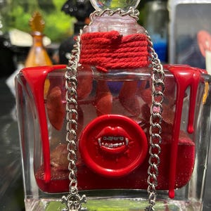 Puede incluir: Una botella de vidrio rectangular llena de l&iacute;quido rojo y purpurina, con gotas de cera roja en las esquinas superiores. Un sello de cera roja con una boca y colmillos est&aacute; centrado en el frente. Una cadena plateada cuelga del cuello.