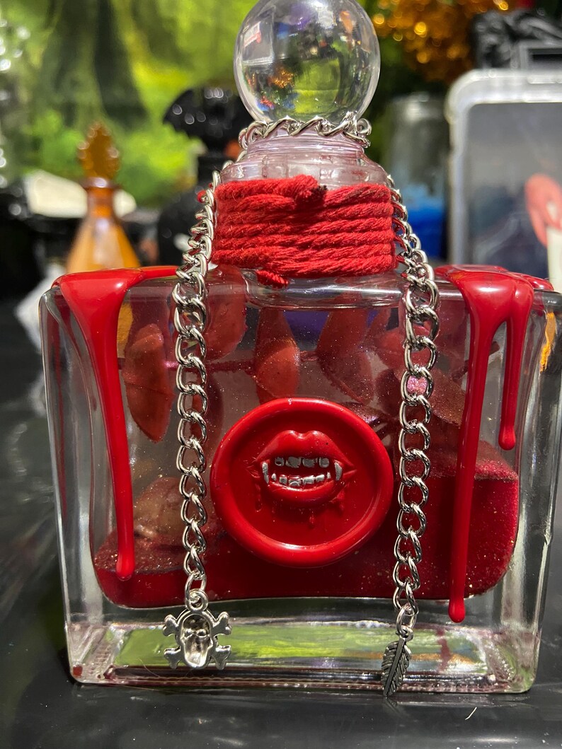 Puede incluir: Una botella de vidrio rectangular llena de l&iacute;quido rojo y purpurina, con gotas de cera roja en las esquinas superiores. Un sello de cera roja con una boca y colmillos est&aacute; centrado en el frente. Una cadena plateada cuelga del cuello.