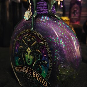 Puede incluir: Una botella de vidrio llena de l&iacute;quido morado y purpurina verde, con una etiqueta que muestra una ilustraci&oacute;n de Medusa de piel verde y el texto Medusa's Braid. El cuello est&aacute; envuelto en hilo morado, cera verde derretida y un dije de cristal.