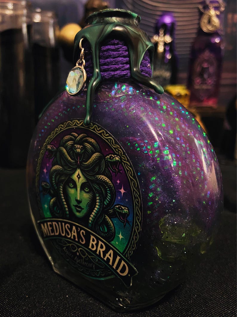 Puede incluir: Una botella de vidrio llena de l&iacute;quido morado y purpurina verde, con una etiqueta que muestra una ilustraci&oacute;n de Medusa de piel verde y el texto Medusa's Braid. El cuello est&aacute; envuelto en hilo morado, cera verde derretida y un dije de cristal.