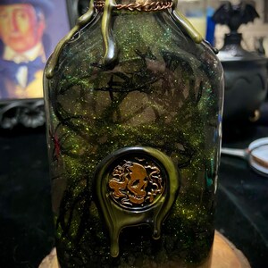 May include: A clear glass bottle filled with green glittery liquid and dark branch-like structures. The neck is wrapped in twine and copper wire, topped with dripping dark wax. A gold-colored circular wax seal featuring a skull is attached.