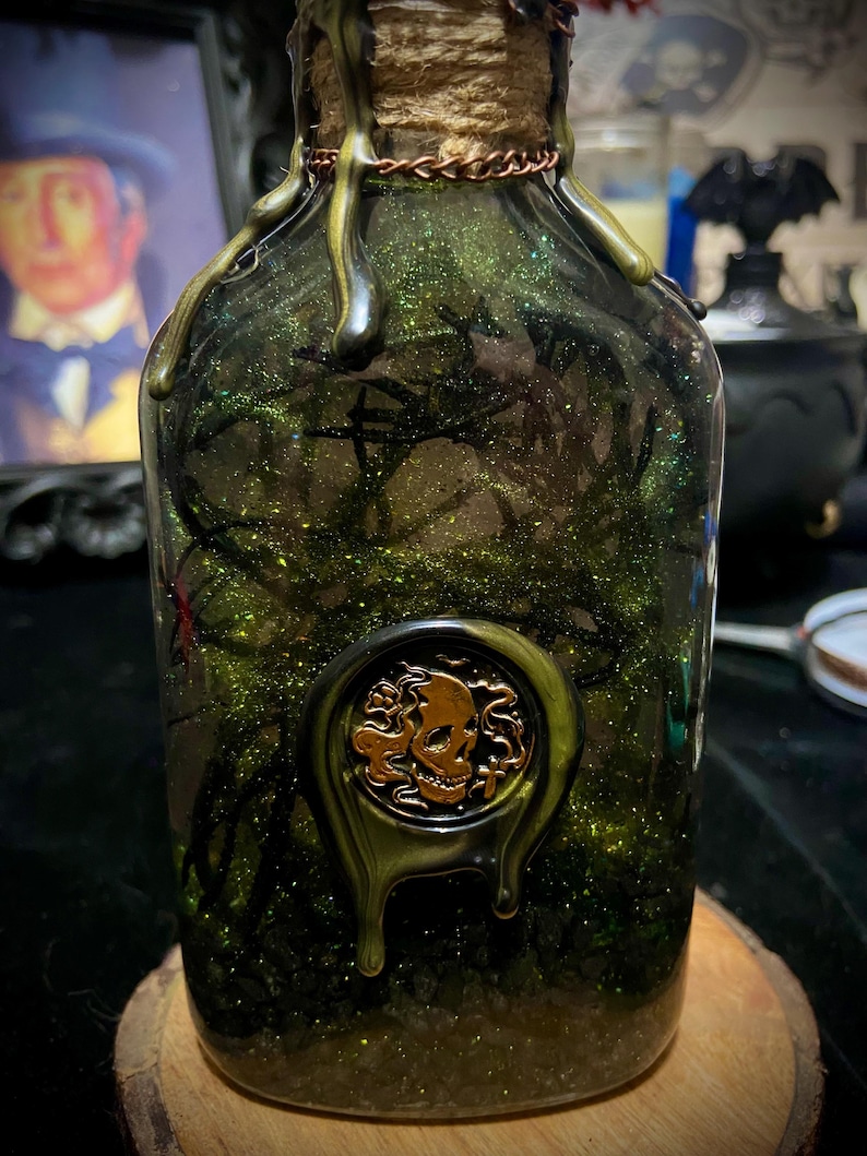 May include: A clear glass bottle filled with green glittery liquid and dark branch-like structures. The neck is wrapped in twine and copper wire, topped with dripping dark wax. A gold-colored circular wax seal featuring a skull is attached.