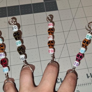 May include: Four finger rings with skull beads in various colors, including red, orange, white, and black. The rings are made of silver-colored wire and are arranged on a grid surface.