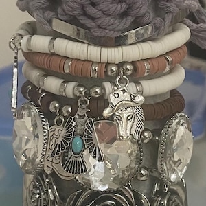 May include: Stacked bracelets in various styles. The top bracelet is pink with the word "Hustle" on it. Other bracelets include a gray macrame, white and brown beaded, and silver charm bracelets with large crystals and rose details.