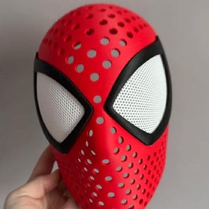 May include: A red full-face mask featuring a perforated surface with numerous circular holes throughout. The mask includes two large, angular eye openings outlined in black, filled with a white, fine-mesh material for visibility.