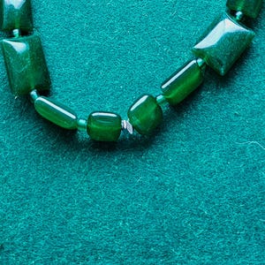 May include: A section of a green beaded necklace. The beads are various shapes, including rectangular and cylindrical, and are strung together. The necklace is set against a teal green background.