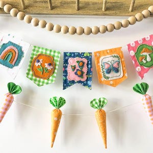 May include: A colorful banner with five fabric pennants, each featuring a different embroidered design: a rainbow, flowers, a butterfly, and a mushroom. Below, are three fabric carrots with green tops. The banner is suspended from a wooden bead garland.