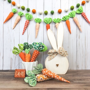 Peut inclure: Un lapin en bois blanc avec un nœud brun et blanc à pois. Le lapin est entouré d'une guirlande de carottes en feutre vert et orange. Une petite pancarte indique "Carottes".