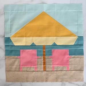 May include: A fabric quilt block depicting a beach scene. The design includes a yellow umbrella, two pink beach chairs, and a light blue sky. The sand is a light brown color.