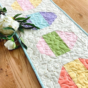 Może przedstawiać: Biały bieżnik stołowy z niebieską lamówką i pikowanym wzorem z pastelowych geometrycznych kształtów. Kształty są ułożone w powtarzający się wzór i przypominają jajka wielkanocne.