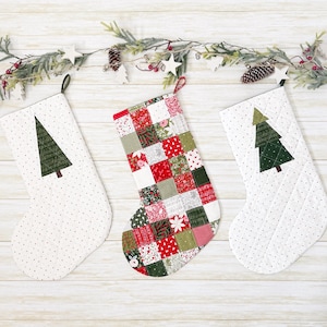May include: Three Christmas stockings. Two white stockings with green tree appliqués and a dotted pattern. The third stocking is a patchwork of red, green, and white fabrics. All have hanging loops and are decorated with a festive garland.