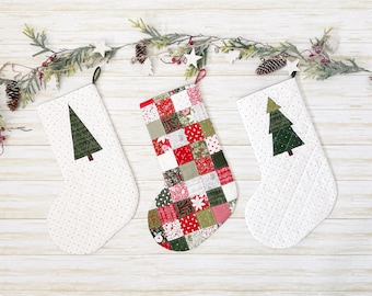 May include: Three Christmas stockings. Two white stockings with green tree appliqués and a dotted pattern. The third stocking is a patchwork of red, green, and white fabrics. All have hanging loops and are decorated with a festive garland.