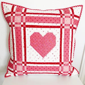 May include: A square decorative pillow with a quilted heart design. The pillow features a white background with small pink hearts, and a large pink heart in the center. Red and pink stripes frame the heart and edges.