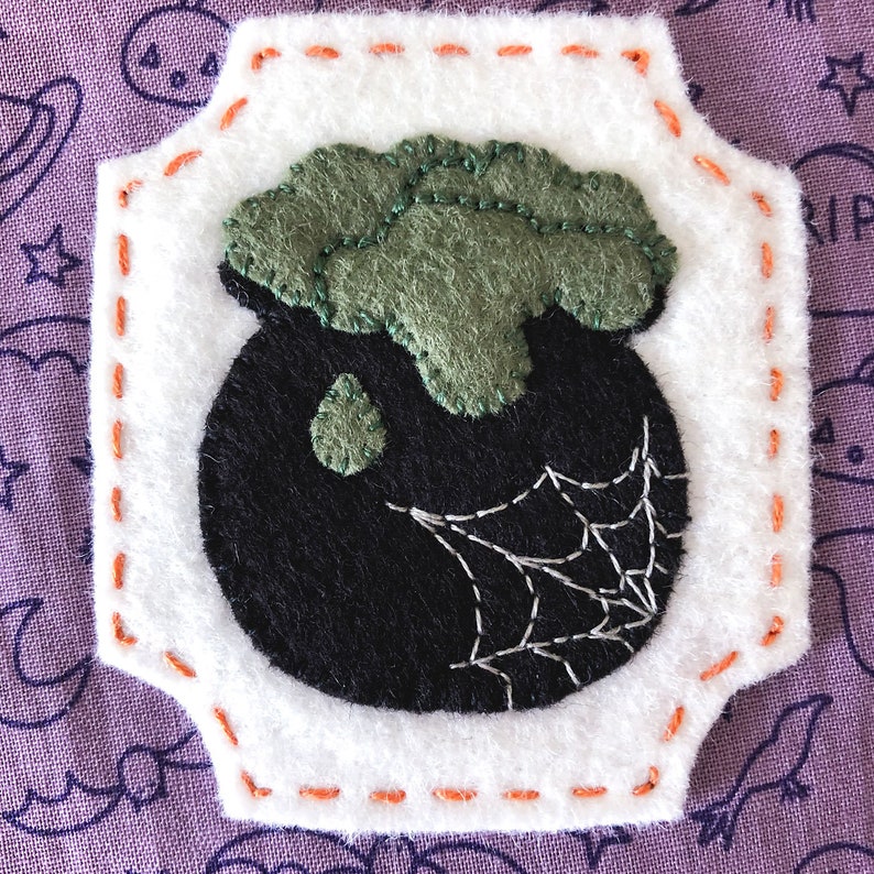 K&ouml;nnte beinhalten: Ein Filz-Halloween-Kessel mit einem gr&uuml;nen Filzdeckel und einem wei&szlig;en Filz-Spinnennetz-Design. Der Kessel ist von einem wei&szlig;en Filzrand mit orangefarbenen N&auml;hten umgeben.