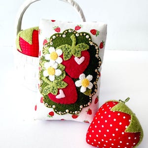 Puede incluir: Un alfiletero rectangular de tela con estampado de fresas, adornado con una aplicación de fieltro de dos fresas rojas, hojas verdes, flores blancas y pequeños corazones rosas, acompañado de dos alfileteros con forma de fresa en una cesta blanca.