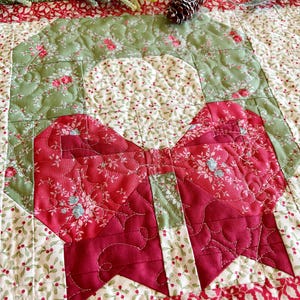May include: A quilted Christmas table runner with a large red bow and a green wreath design. The bow and wreath are made of floral fabric, and the background is cream with red berry accents. A pinecone and greenery are on top.