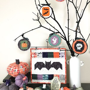 K&ouml;nnte beinhalten: Ein Halloween-Quilt mit einem schwarzen Fledermaus-Design, umgeben von handgefertigten Stoffk&uuml;rbissen, Grabsteinen und Ornamenten. Die Ornamente zeigen einen Geist, einen Sch&auml;del, einen Kessel und Flederm&auml;use.