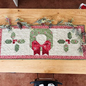 May include: A rectangular Christmas table runner with a central wreath design and holly accents. The runner features a red and green color scheme, with a red bow on the wreath. Pinecones and greenery decorate the wooden table.