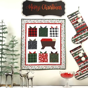 May include: A white quilted wall hanging with a Christmas theme. The quilt features a manger scene with a brown wooden manger and a red blanket. There are also six presents wrapped in different patterns, including plaid, black and white, and green. The quilt is surrounded by a white border with a red and green floral pattern. The quilt is hanging on a white wall with a black sign that says "Merry Christmas" above it. There are also two Christmas stockings hanging on the wall.