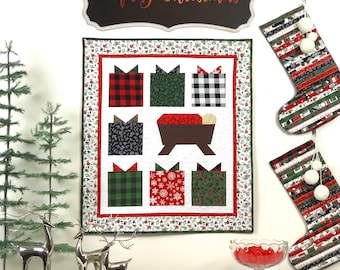 Peut inclure: Une courtepointe murale blanche sur le thème de Noël. La courtepointe représente une scène de crèche avec une crèche en bois brun et une couverture rouge. Il y a aussi six cadeaux emballés dans différents motifs, dont du plaid, du noir et blanc et du vert. La courtepointe est entourée d'une bordure blanche avec un motif floral rouge et vert. La courtepointe est accrochée à un mur blanc avec une pancarte noire qui dit "Joyeux Noël" au-dessus. Il y a aussi deux chaussettes de Noël accrochées au mur.