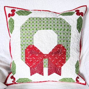 May include: Square quilted pillow with a Christmas wreath design. The wreath features a green plaid center, a red bow, and green leaves with red berries. The pillow has a white background with a red border.