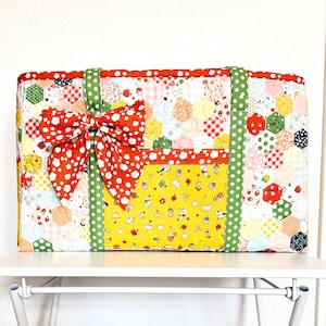 May include: A rectangular quilted tote bag featuring a patchwork of hexagonal fabric patterns in various colors. The bag includes two green polka-dot straps, a large red floral bow on the front, and a yellow pocket with small fruit icons.