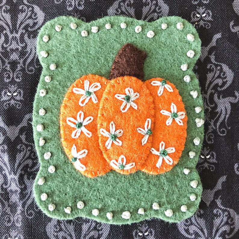 K&ouml;nnte beinhalten: Eine Filz-Applikation in Form eines K&uuml;rbisses auf gr&uuml;nem Hintergrund mit wei&szlig;en G&auml;nsebl&uuml;mchen-Stickereien. Der K&uuml;rbis ist orange mit einem braunen Stiel.