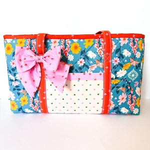 Puede incluir: Un bolso tote acolchado con fondo azul cerceta y un estampado colorido de flores y mariposas. El bolso tiene asas naranjas, un lazo rosa y un bolsillo frontal de tela blanca con pequeños patrones de corazones verdes y rosas.