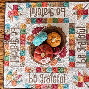May include: A square fabric quilt featuring a patchwork border of colorful squares and the text be grateful printed on each side. A dark wooden bowl sits in the center, containing three fabric pumpkins in orange, red, and blue, plus fabric leaves.