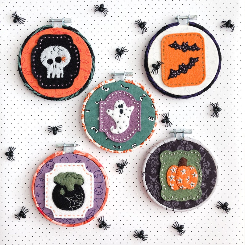 K&ouml;nnte beinhalten: F&uuml;nf Halloween-Motive aus Filz mit Stickerei. Jedes Ornament ist ein runder, wei&szlig;er Filzkreis mit einem schwarzen Filzrand und einem anderen Halloween-Motiv. Die Motive umfassen einen Sch&auml;del, ein Gespenst, einen K&uuml;rbis, eine Fledermaus und einen Kessel.