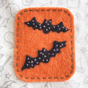 K&ouml;nnte beinhalten: Zwei schwarze Filzflederm&auml;use mit lila N&auml;hten auf einem orangefarbenen Filzhintergrund. Die Flederm&auml;use sind mit gr&uuml;nem Garn um die R&auml;nder gen&auml;ht.