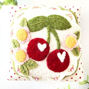 Puede incluir: Un alfiletero de tela cuadrado con fondo blanco y lunares rojos. El centro muestra dos cerezas de fieltro rojo con detalles en forma de corazón blanco, rodeadas de hojas de fieltro verde y flores de fieltro rosas y amarillas con lentejuelas.